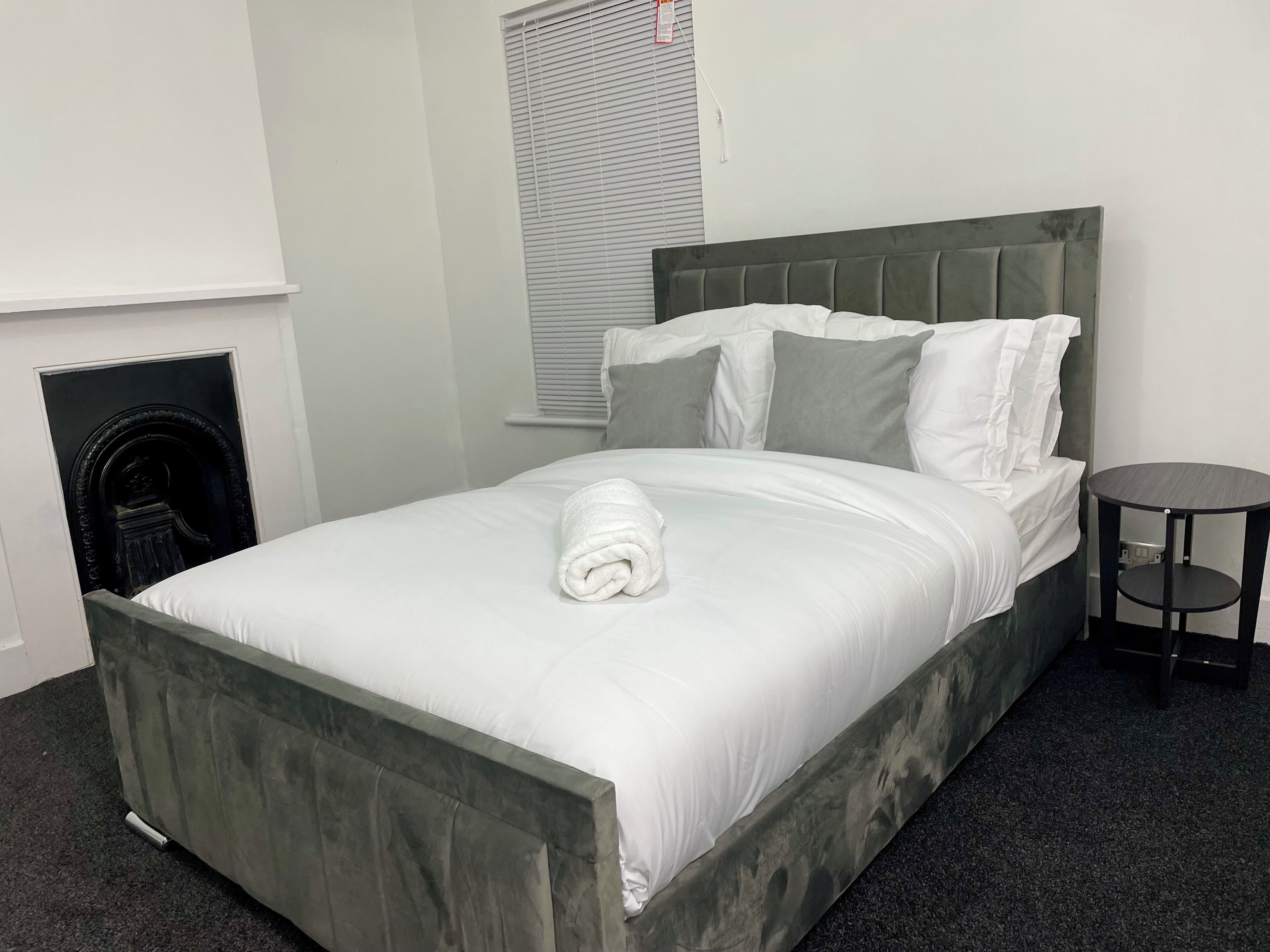 A neatly made bed with white linens and a rolled towel, a small round table beside it, and a fireplace in the corner.