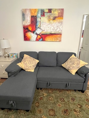 A gray sectional sofa with two decorative pillows, a lamp, and a colorful abstract painting on the wall. - Cozy Suite #105 (Philadelphia)