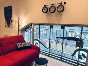 A red sofa with patterned cushions, a black round ottoman, and a wall-mounted clock.