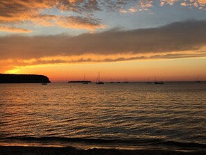 A sunset over a calm sea with sailboats and a distant landmass.