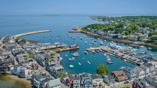 Un pueblo costero con un puerto deportivo lleno de barcos, rodeado de edificios residenciales y una amplia zona verde.