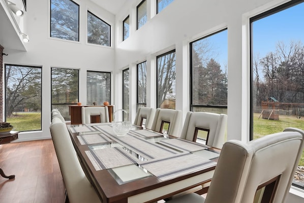 A modern dining room with a long table, multiple chairs, and large windows offering a view of the outdoors.