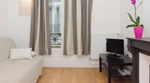 A living room with a sofa, a TV on a stand, a potted plant, and a window with curtains.