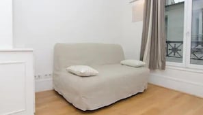 A plain beige sofa with two pillows, a window with curtains, and a wooden floor.