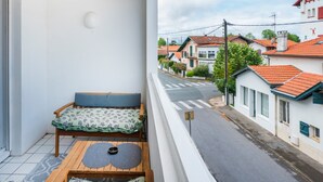 A balcony with a wooden table, a cushioned bench, and a view of a street with houses. - Apartment 'Residence Le Sabaou' with Balcony (Biarritz)