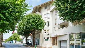 A street with a row of white buildings, trees lining the road, and parked cars. - Apartment 'Residence Le Sabaou' with Balcony (Biarritz)