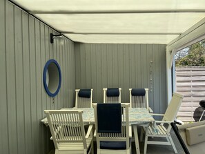 Een overdekt terras met een tafel en vier stoelen, een ronde blauwe spiegel aan de muur, en een houten hek op de achtergrond.