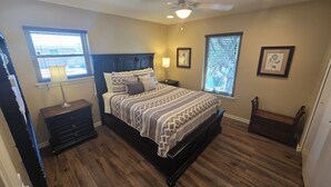A bedroom with a wooden floor, a bed with patterned bedding, a nightstand, a lamp, and two windows with blinds. - Clean, Comfortable Home Away From Home! (El Paso)