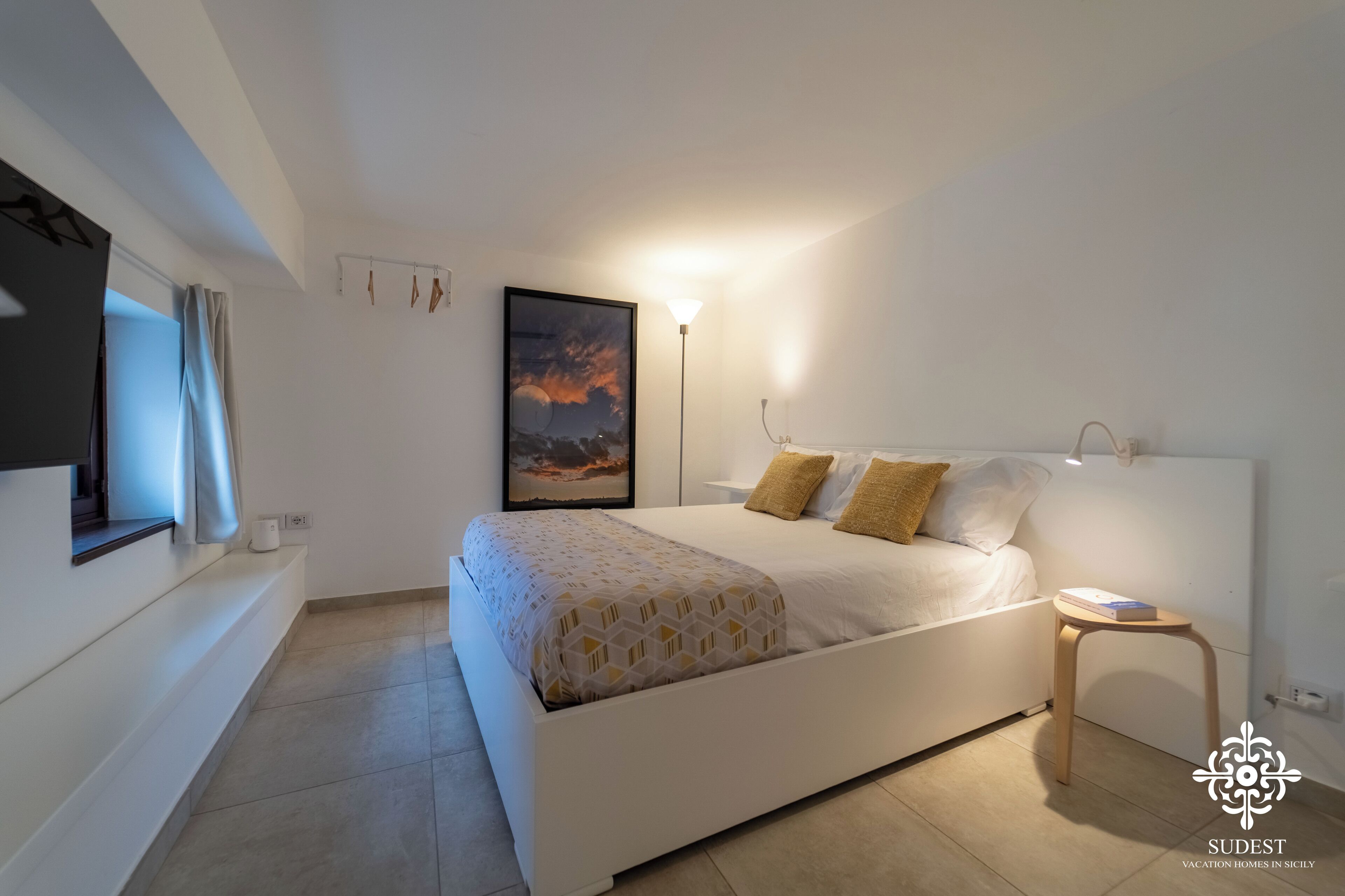 A modern bedroom with a large bed, a bedside table, a wall-mounted TV, and a framed picture.