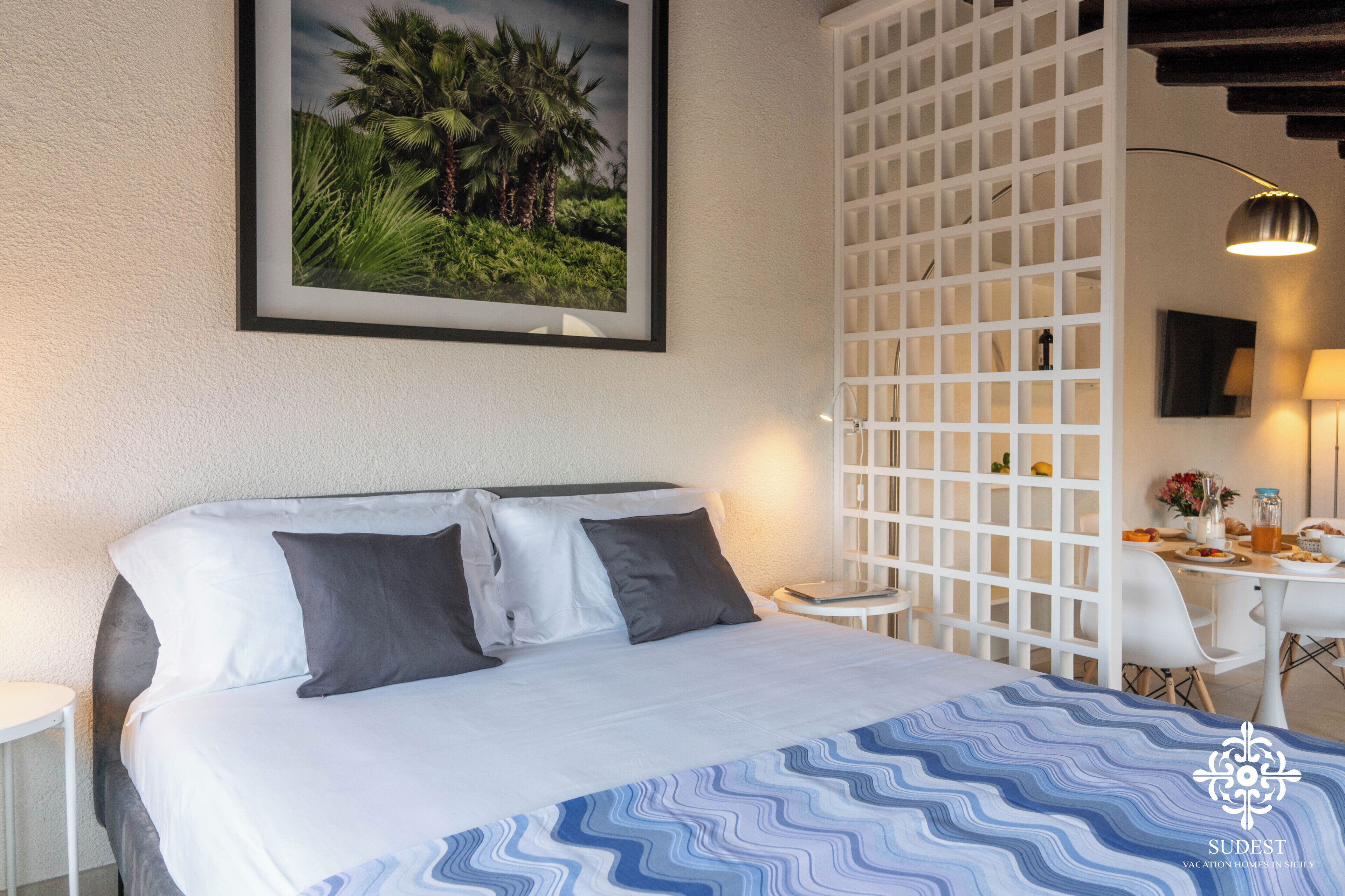 Ein Hotelzimmer mit einem Bett, einer weißen Trennwand, einem kleinen Tisch mit einer Lampe und einem gerahmten Bild an der Wand.