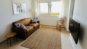 A living room with a brown leather sofa, a small wooden table, a woven rug, a wicker chair, and a flat-screen TV. - Mundet 1 Baix Mar CostaBravaSi - Sea, beach and AC (Sant Antoni de Calonge)