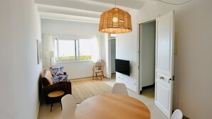 A modern living room with a round wooden table, white chairs, a wicker pendant light, a TV mounted on the wall, and a sofa with patterned cushions. - Mundet 1 Baix Mar CostaBravaSi - Sea, beach and AC (Sant Antoni de Calonge)