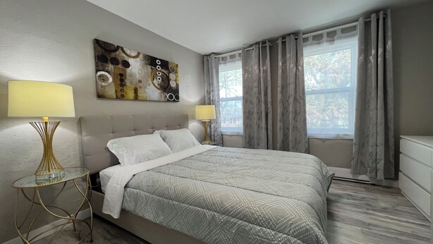Un dormitorio moderno con una cama grande, dos mesitas de noche con lámparas, una lámpara de techo, una ventana con cortinas y una obra de arte en la pared.