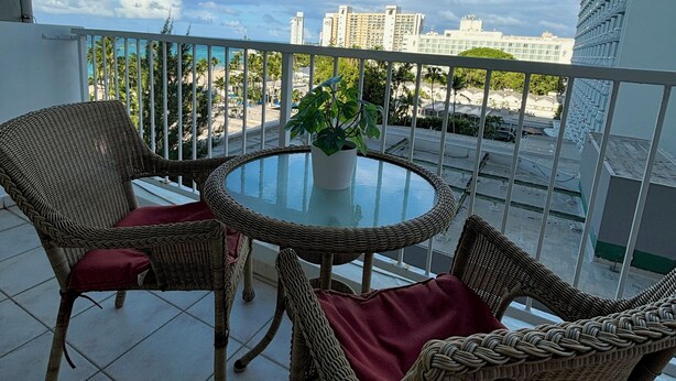 A balcony with wicker furniture and a glass table, overlooking a beach and buildings.