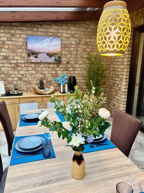 A dining area with a brick wall, a painting, and a table set with blue placemats, plates, and a vase of flowers.