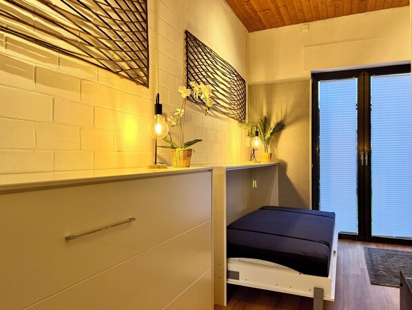 Ein gemütliches Schlafzimmer mit einem Bett, einer kleinen Ablage mit Pflanzen, einer Wandleuchte und einer Schiebetür zum Balkon.