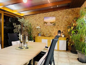 A dining area with a wooden table, chairs, and a brick wall with a painting. There are potted plants and a decorative vase on the wall.