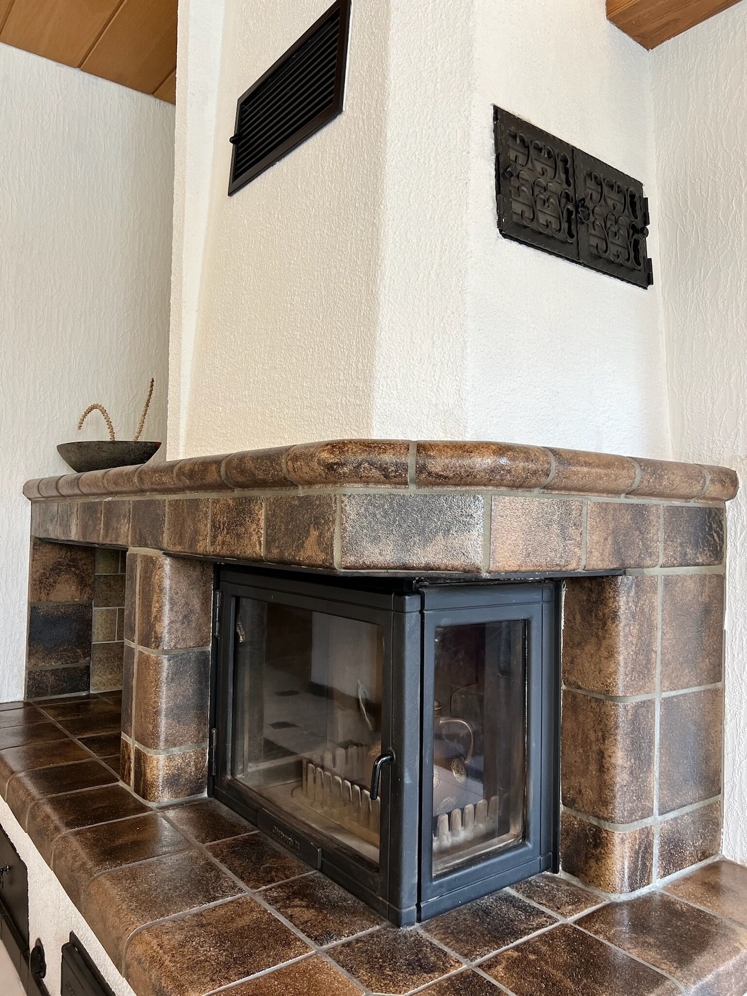 A stone fireplace with a glass door, set in a corner with a white wall and a wooden ceiling.