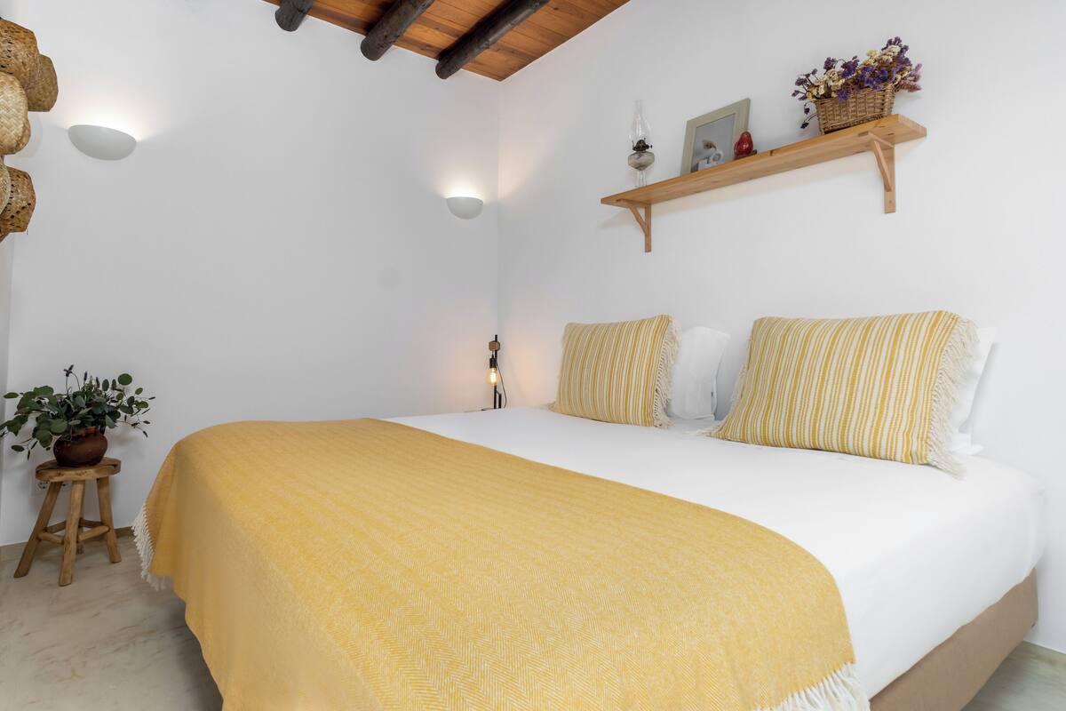 A bedroom with a large bed, a wooden headboard, a small stool with a plant, and a shelf with flowers and a lamp.