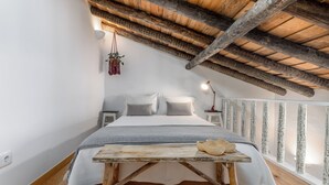 A bedroom with a wooden ceiling, a bed with white linens, a small table with shoes, and a hanging plant.