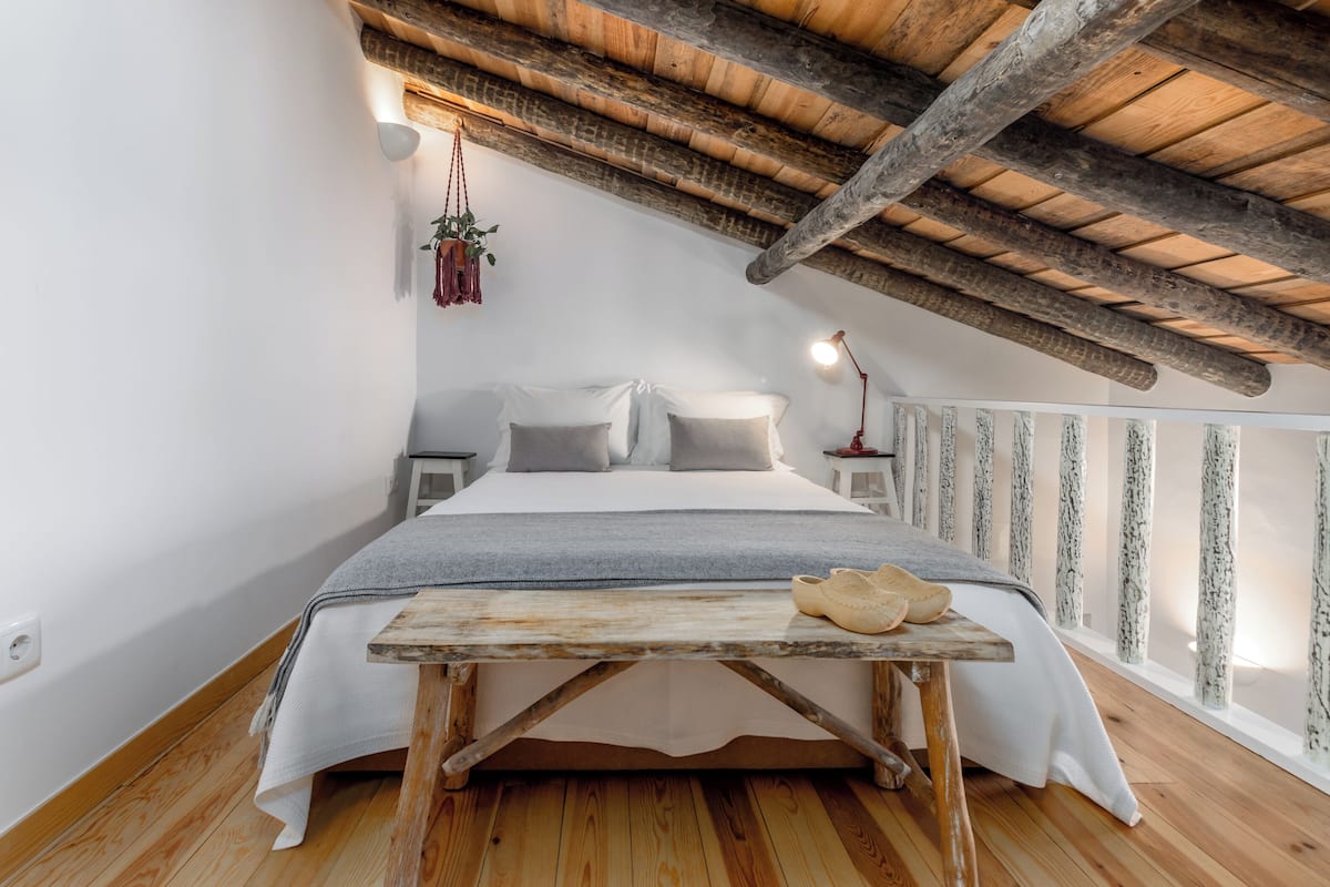 A bedroom with a wooden ceiling, a bed with white linens, a small table with shoes, and a hanging plant.