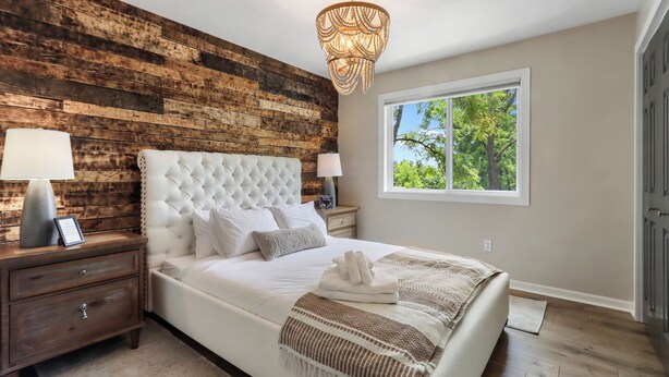 Een slaapkamer met een houten hoofdeinde, een groot raam met uitzicht op bomen, een kroonluchter en een net opgemaakt bed met handdoeken.