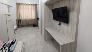 A modern room with a flat-screen TV mounted on a wooden wall, a small kitchenette, and a bathroom with a toilet and shower.