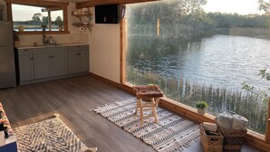 Una cocina moderna con un ventanal grande que da a un lago, vigas de madera y una zona de estar acogedora con un tapete y un taburete.