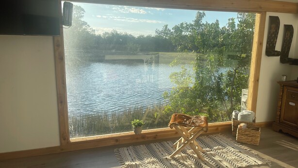 Una habitación con un ventanal grande que da a un lago, una silla de madera, un tapete tejido y un armario de madera.