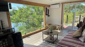 Una habitación amplia con un gran ventanal con vistas al agua, una zona de estar acogedora con un sofá estampado, una pequeña mesa redonda con un jarrón y un mueble de madera con tazas.
