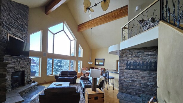 A spacious living room with a stone fireplace, large windows, and a high ceiling with exposed beams. - Charming Big Sky meadows house. Trails and Town Center a short walk away. (Big Sky)