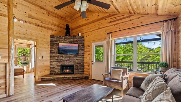 A cozy cabin interior with a stone fireplace, wooden floors, a ceiling fan, and a large window with a view of greenery.