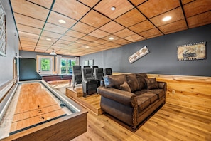 Une table de billard, un canapé en cuir marron et plusieurs chaises noires dans une pièce aux murs lambrissés et dont un des murs est peint en gris.