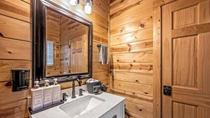 Une salle de bain aux murs lambrissés, dotée d’un grand miroir, d’un lavabo et de trois flacons de produits de toilette.