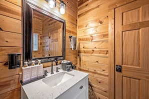 Une salle de bain aux murs lambrissés, dotée d’un grand miroir, d’un lavabo et de trois flacons de produits de toilette.
