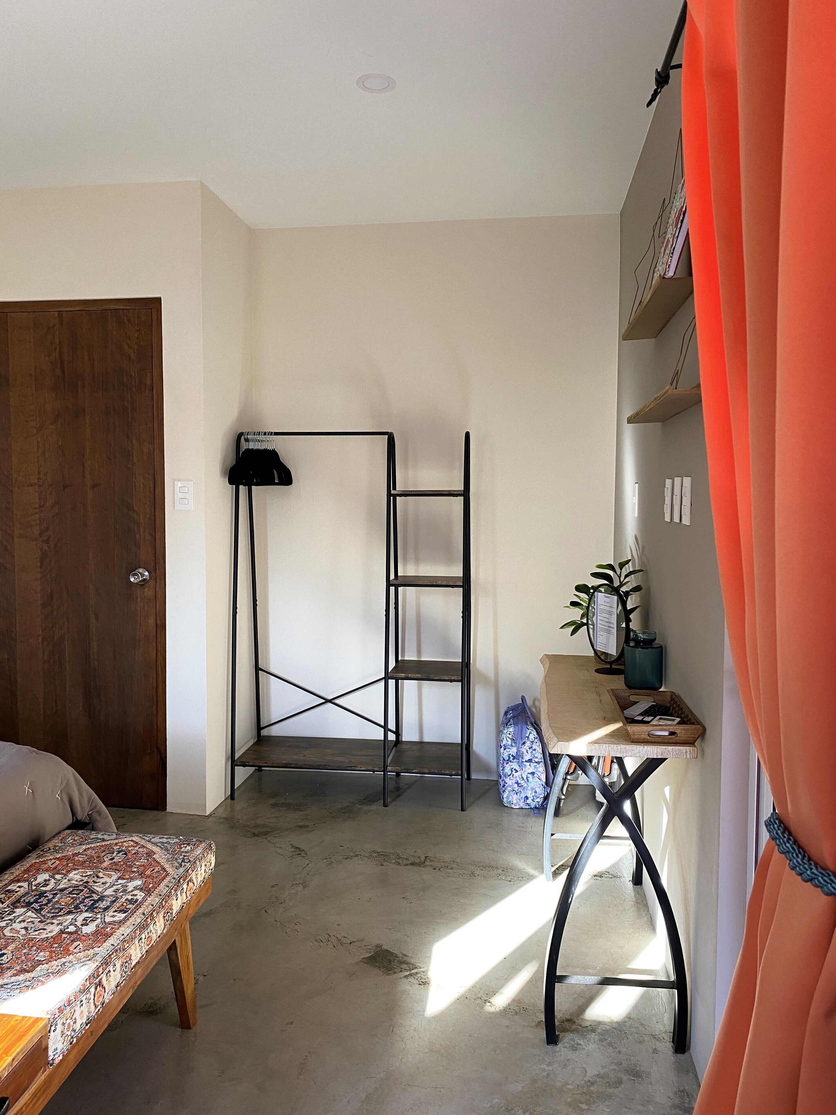 Une pièce avec une porte en bois, un lit recouvert d’une couvre-lit à motifs, une étagère en métal, un bureau avec une chaise et une plante en pot.