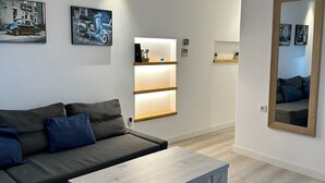 Un salon moderne avec un canapé gris, une table basse en bois et un ventilateur de plafond. On y trouve des étagères murales ornées d’objets décoratifs et un miroir.