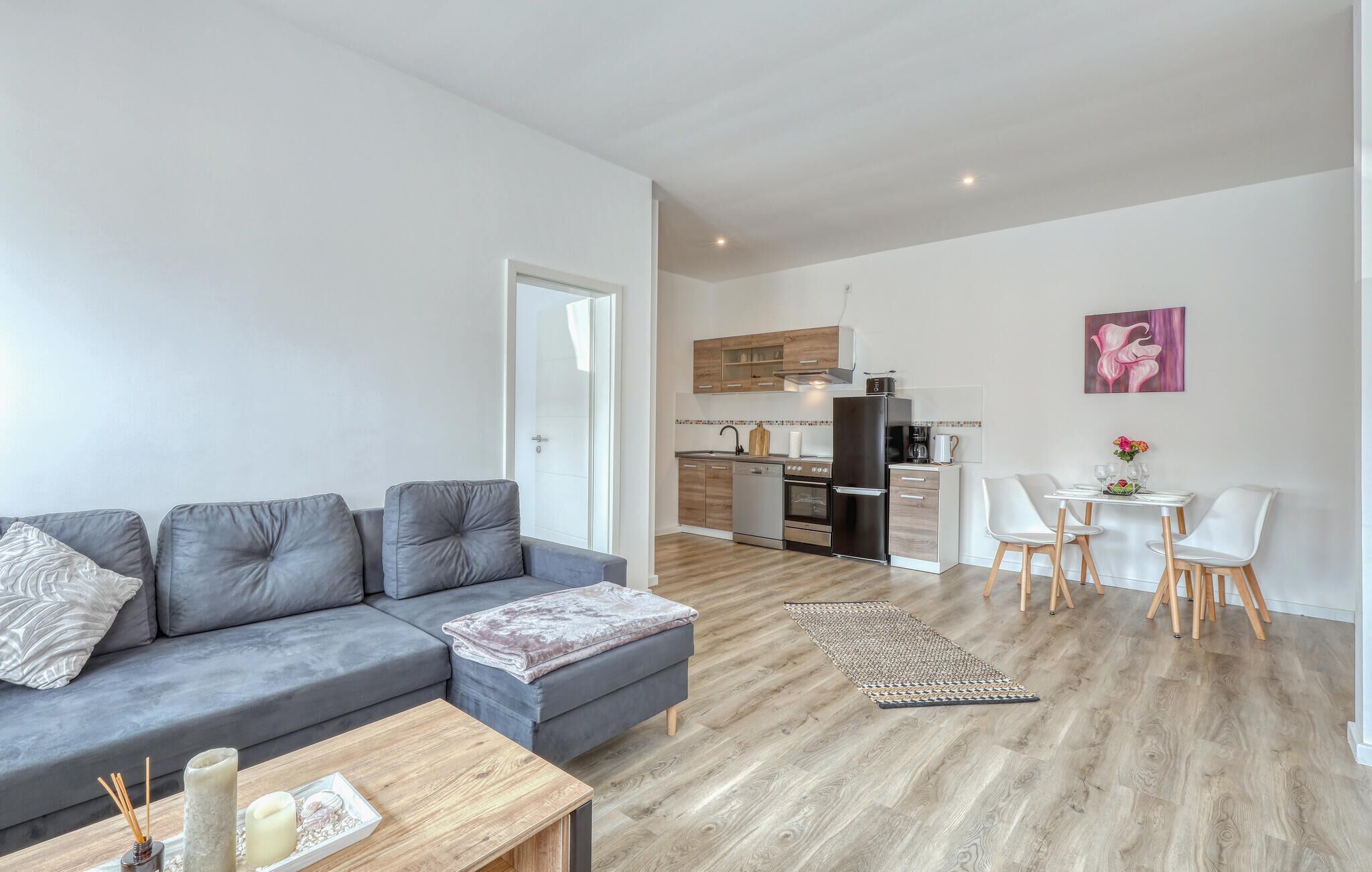 A modern apartment with a grey sofa, a wooden coffee table, a kitchen with wooden cabinets, and a dining area with white chairs.