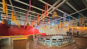 Un espacio interior amplio con una mesa redonda central rodeada de sillas, un mostrador de ladrillo y decoraciones colgantes coloridas.