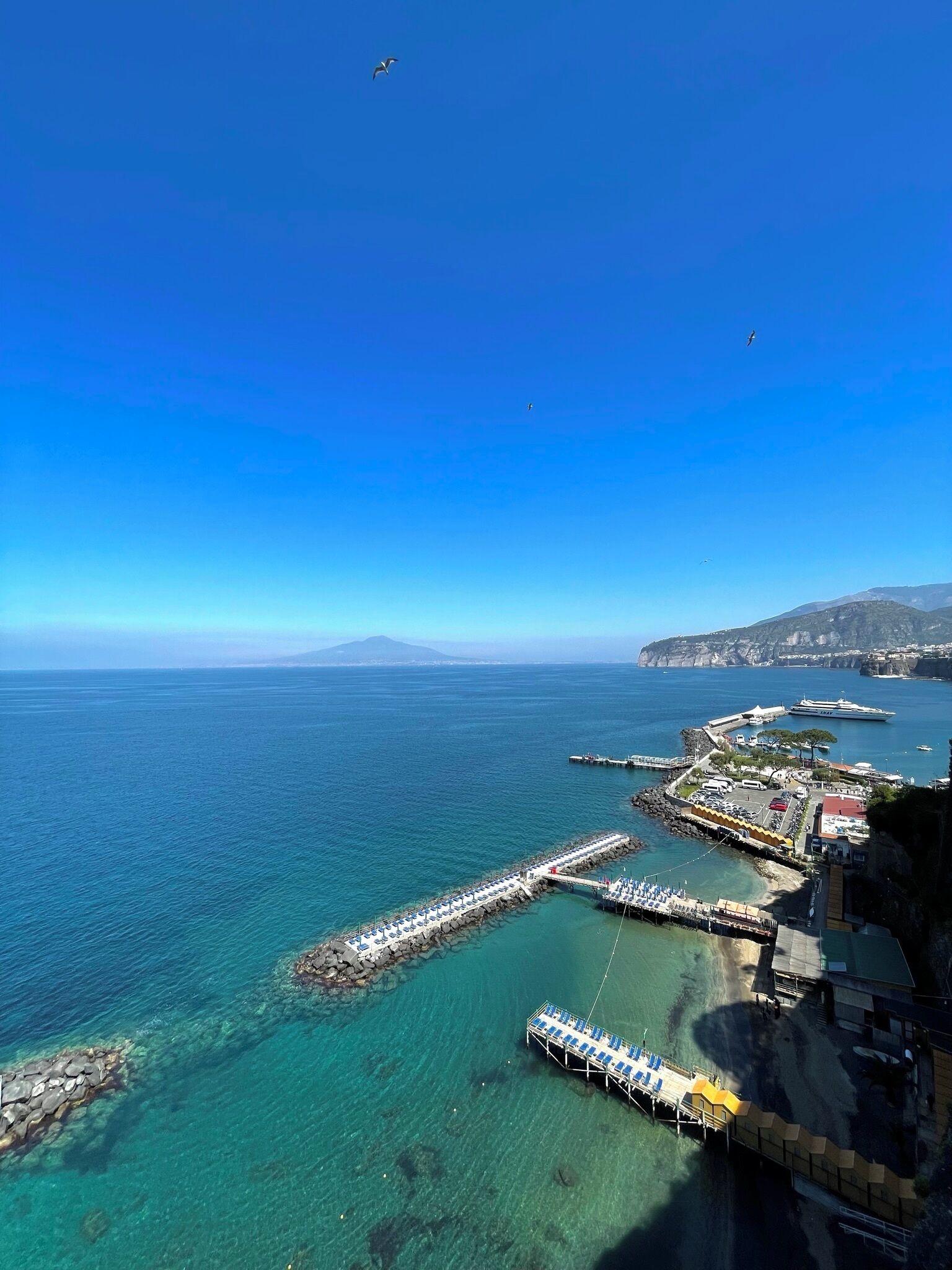 A coastal pier with clear blue waters, a dock with blue chairs, and a building with a green roof.