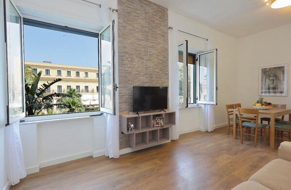 A modern living room with a flat-screen TV, wooden flooring, a stone accent wall, and large windows offering a view of a building and greenery outside.