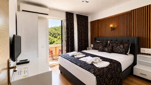A hotel room with a bed, a television, a desk, and a balcony with a view of trees. - Olive Branch Hotel (Ayvalik)