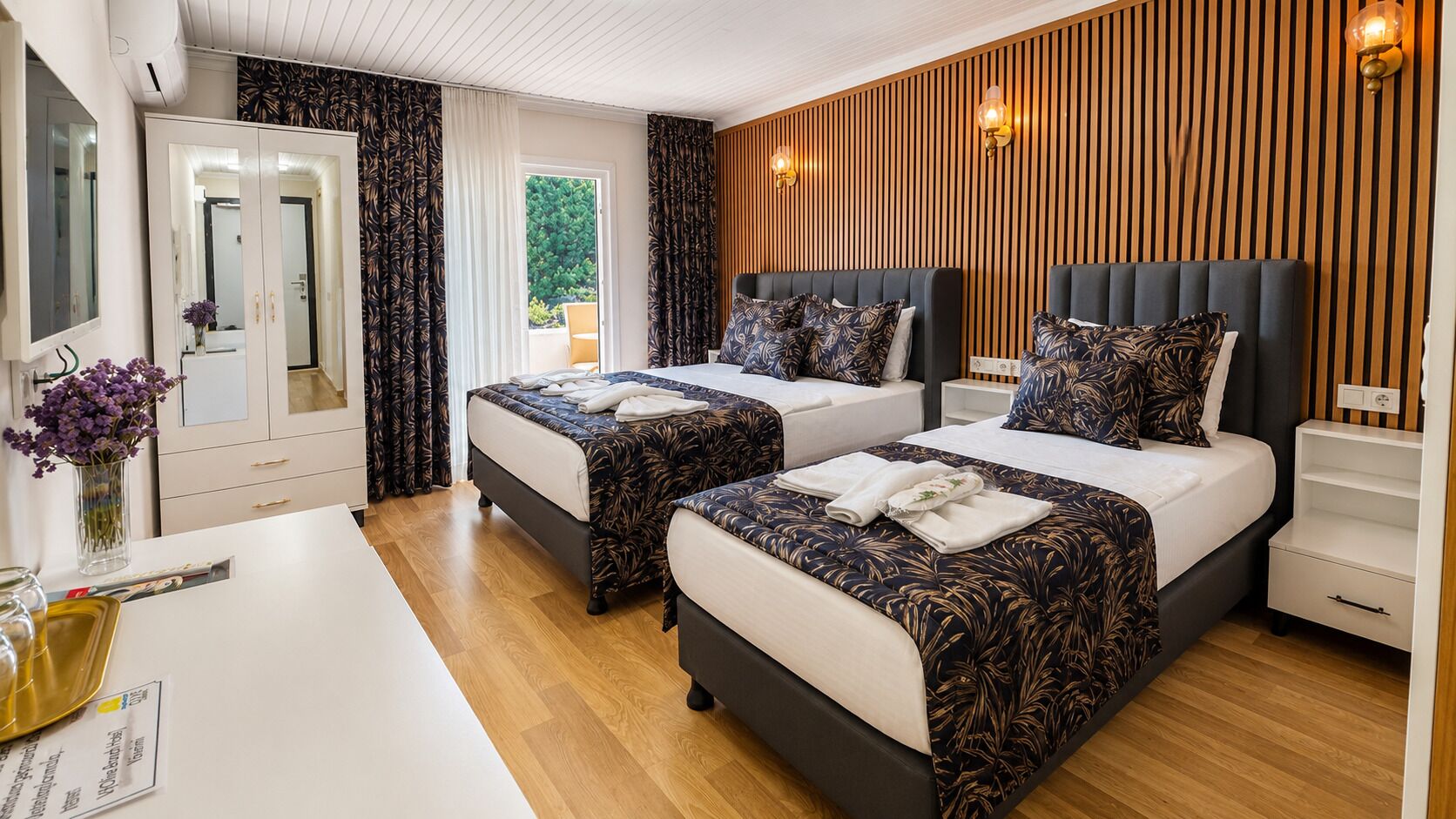 A hotel room with two beds, a white desk, a mirror, a wooden wall panel, and a window with curtains.
