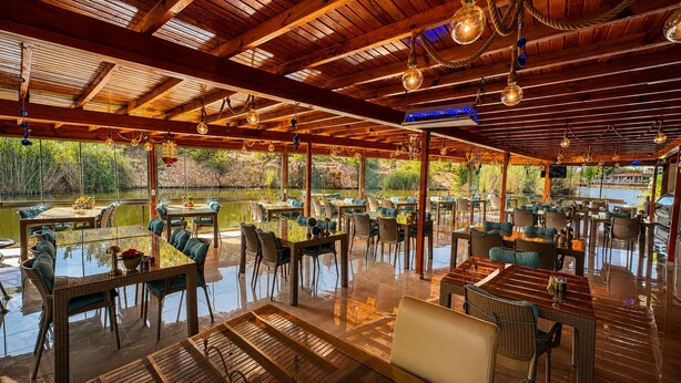 A restaurant with outdoor seating overlooking a body of water, featuring wooden beams and hanging light fixtures. - Olive Branch Hotel (Ayvalik)