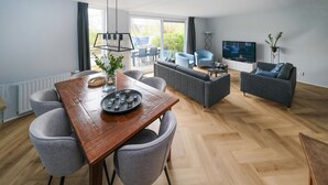A modern living room with a wooden dining table, grey upholstered chairs, a grey sofa set, a flat-screen TV, and a large window offering a view of the outdoors.