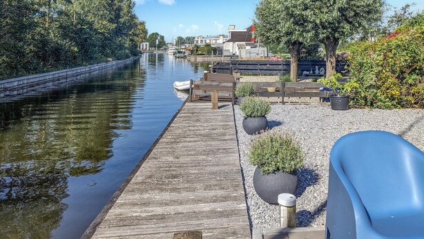 A wooden dock extending into a calm river, with boats docked on the left. There are potted plants and a bench along the dock.