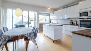 A modern kitchen with white cabinets, a built-in oven, and a microwave. There's a dining area with a table and chairs, and a large window providing natural light.