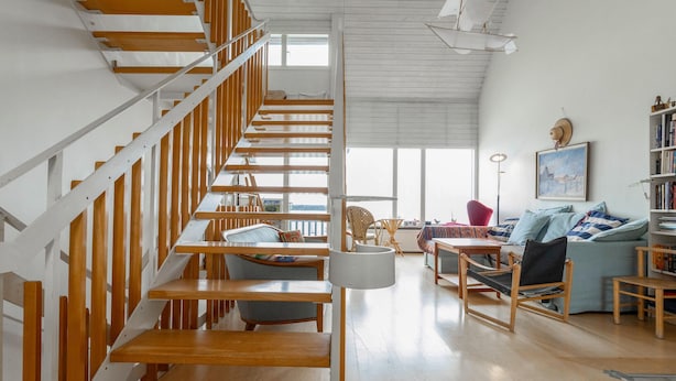 A spacious living area with a wooden staircase, a sofa set, a dining table, and a bookshelf.