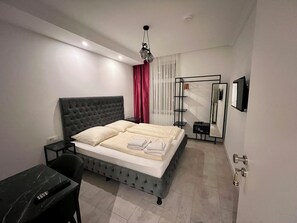 A modern bedroom with a tufted headboard bed, a bedside table, a desk with a chair, a mirror, and a wardrobe. - Durmaz Hotel (Hannover)