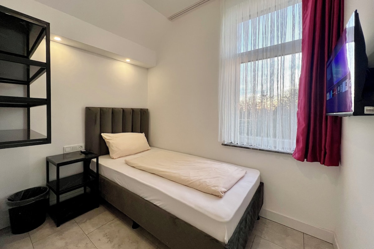 A single bed with a headboard, a small bedside table, a trash bin, a window with sheer curtains, and a flat-screen TV mounted on the wall.